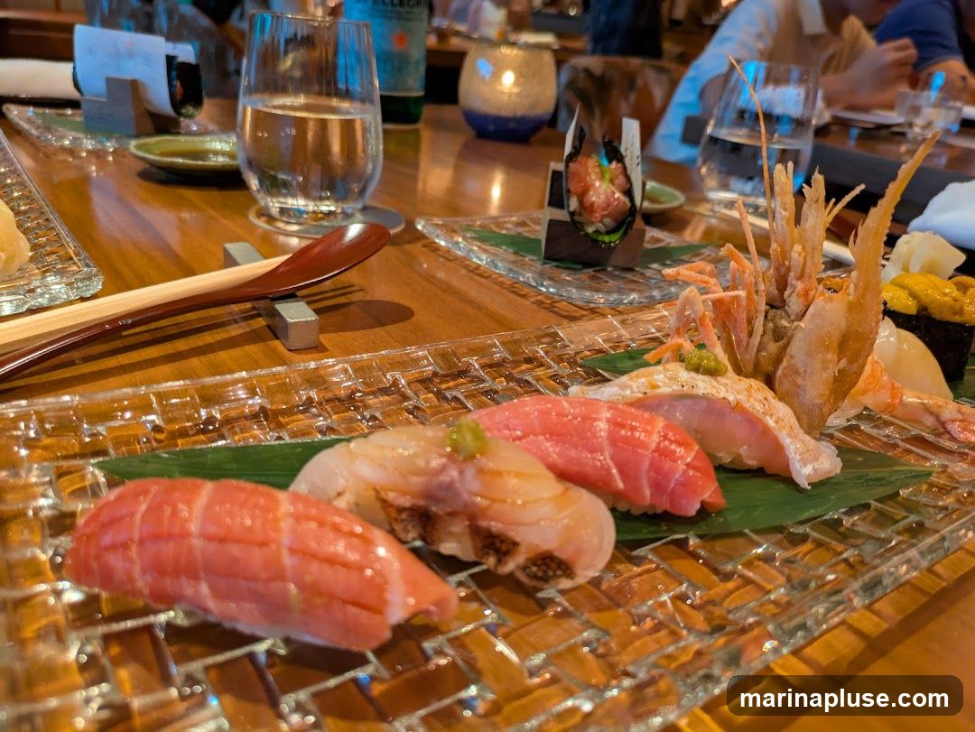A close-up shot of a sushi platter featuring various types of nigiri and a tempura shrimp. The sushi includes what appears to be tuna, salmon, and sea urchin. The platter is made of clear, textured glass and is set on a wooden table. In the background, there are glasses of water, a bottle of Pellegrino, and a lit candle.