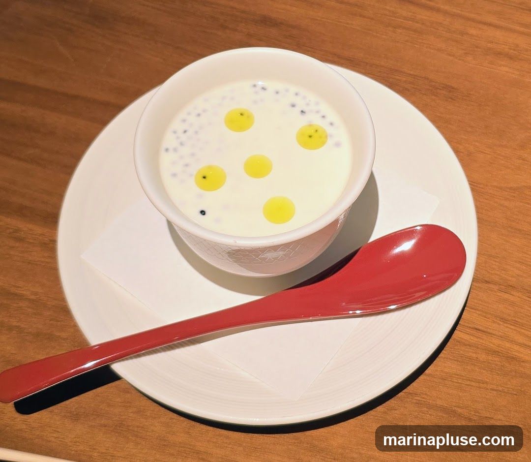A white bowl filled with a creamy white dessert, topped with small dark spheres and several bright yellow oil droplets. The bowl is placed on a white saucer with a white paper liner. A red spoon rests on the saucer. The setting appears to be a wooden table.