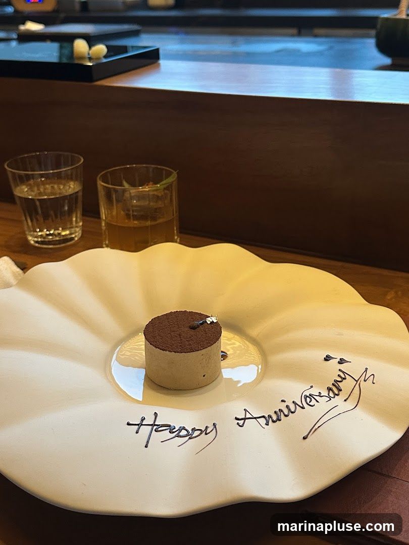 A small, cylindrical dessert dusted with cocoa powder and topped with a tiny gold leaf sits on a decorative white plate. The plate has a scalloped edge and is inscribed with the words "Happy Anniversary" in dark, elegant script. Two glasses, one with a clear liquid and the other with an amber liquid and ice, are visible in the background. The setting appears to be a restaurant or dining establishment.