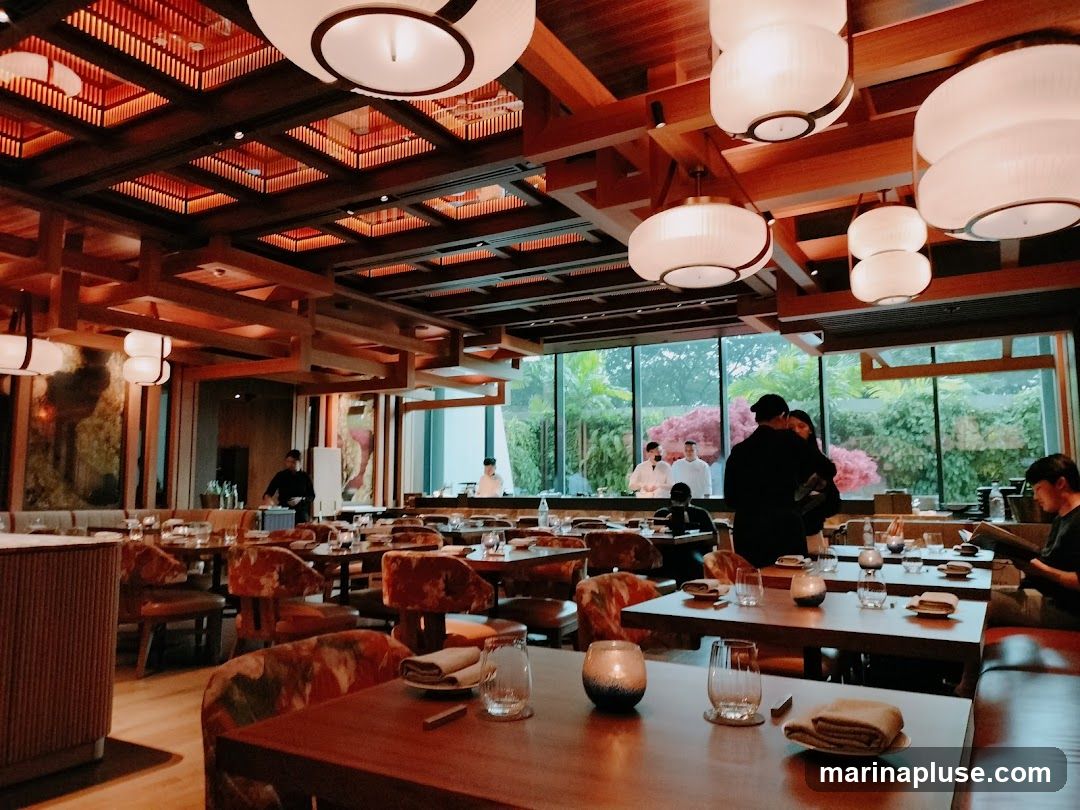 The interior of a modern restaurant with wooden accents, ambient lighting, and tables set for dining. Staff members are visible in the background, and diners are seated at some tables. Large windows offer a view of greenery outside.