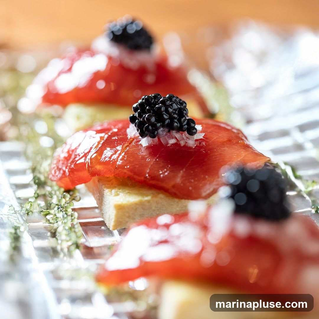 A close-up, shallow depth of field shot showcases three canapés arranged on a clear, textured platter. Each canapé consists of a rectangular piece of bread topped with thinly sliced smoked salmon, finely diced white onion, and a dollop of black caviar. Delicate green sprigs are scattered around the canapés, adding a touch of freshness. The background is softly blurred, with warm, golden tones suggesting ambient lighting.