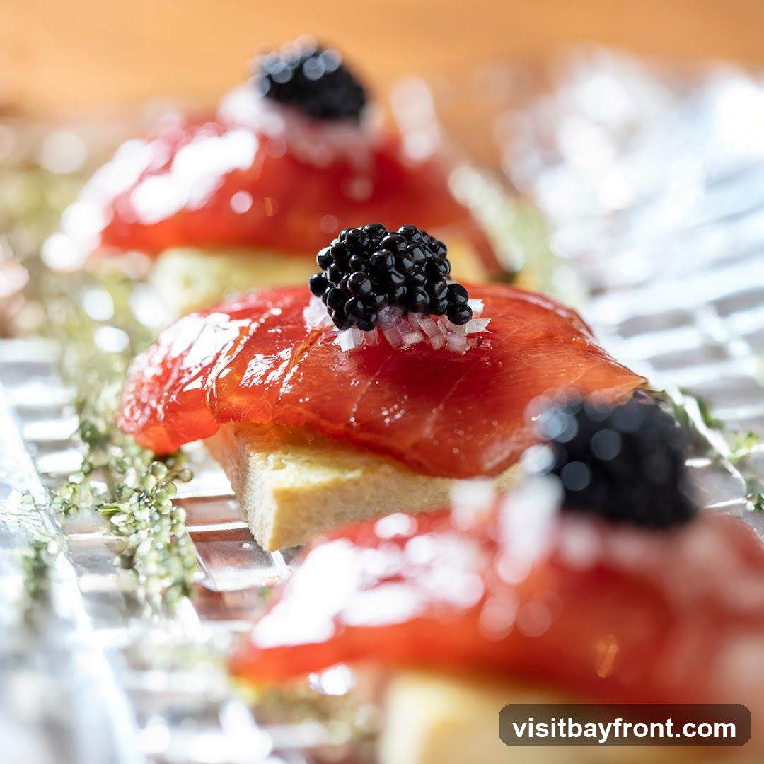 A close-up, shallow depth of field shot showcases three canapés arranged on a clear, textured platter. Each canapé consists of a rectangular piece of bread topped with thinly sliced smoked salmon, finely diced white onion, and a dollop of black caviar. Delicate green sprigs are scattered around the canapés, adding a touch of freshness. The background is softly blurred, with warm, golden tones suggesting ambient lighting.