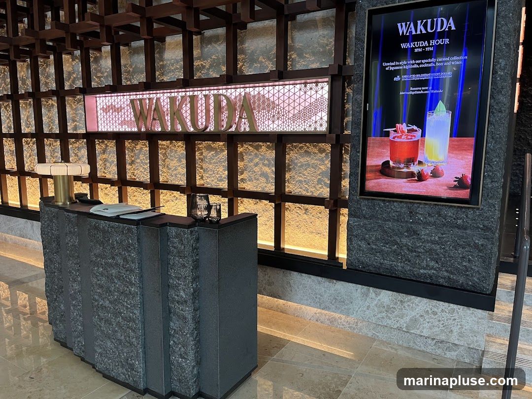 A reception desk with a lamp and glasses in front of a wall with a large illuminated sign that says WAKUDA. To the right, a digital screen displays an advertisement for Wakuda Hour, featuring cocktails and the text "Unwind in style with our specially curated collection of Japanese highballs, cocktails, beer and wines."