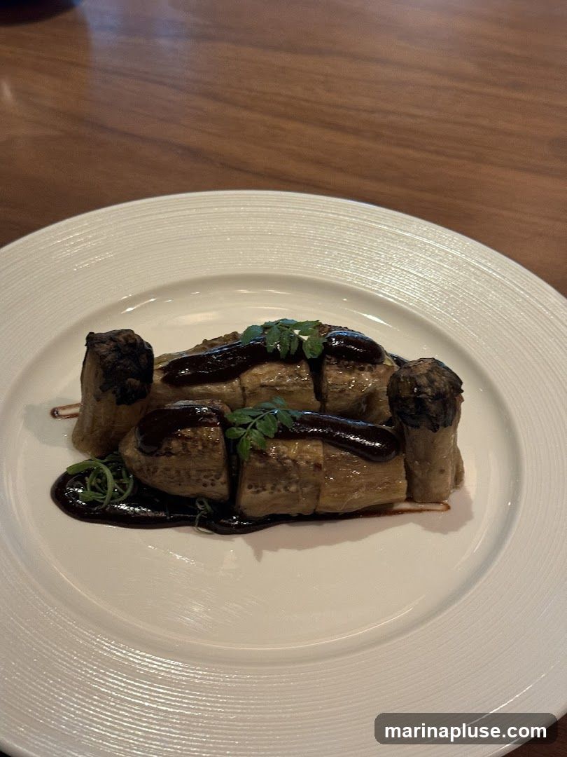 A close-up shot of a dish featuring sliced eggplant wrapped in a dark sauce, garnished with herbs, served on a white plate. The plate is on a wooden table.