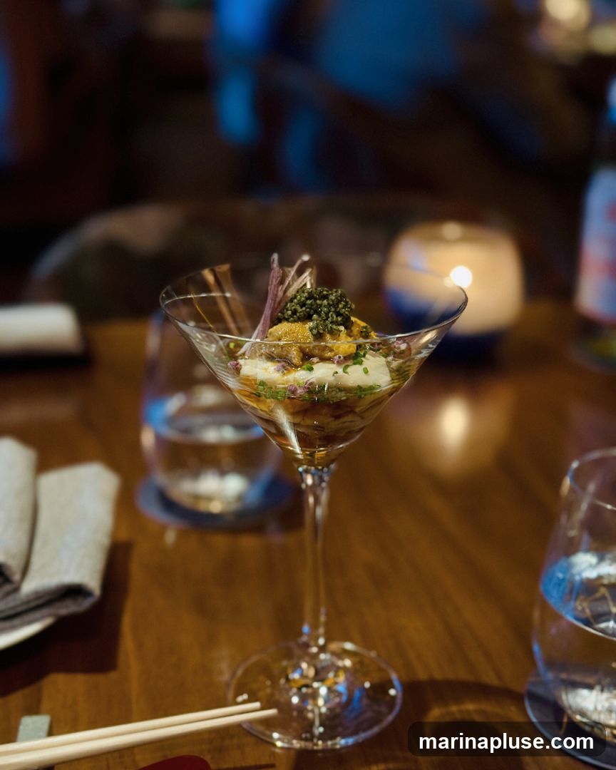 A close-up shot of a delicate appetizer served in a martini glass. The dish features layers of seafood, possibly uni and caviar, garnished with herbs and edible flowers. The setting appears to be a dimly lit restaurant with a candle and glasses of water on a wooden table.