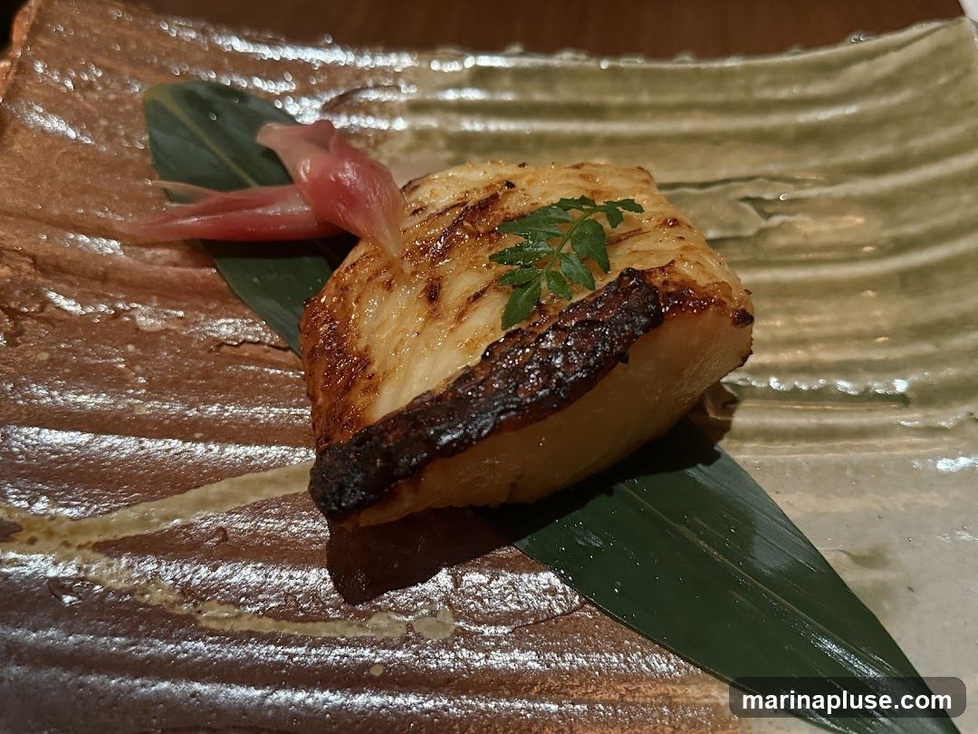 A close-up shot of a piece of grilled fish, served on a textured ceramic plate with a green leaf and pickled ginger. The fish appears to be flaky and well-seasoned, with a slightly charred exterior.