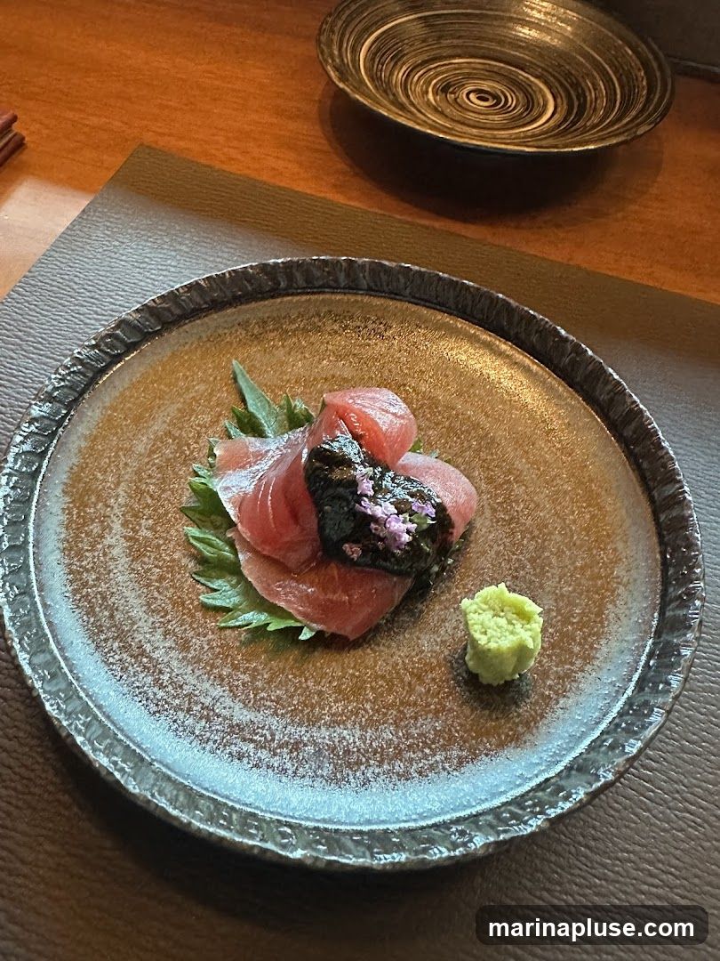 A close-up shot of a sashimi appetizer served on a textured, dark-rimmed plate. The dish features several slices of red tuna, artfully arranged on a bed of green leaves. A dollop of dark, possibly seasoned paste, is placed on top of the tuna, garnished with delicate purple flowers. To the side, a small mound of bright green wasabi completes the presentation. The plate sits on a dark placemat on a wooden table. Another decorative plate with a spiral pattern is visible in the background.