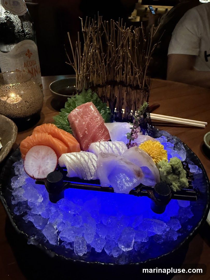 A close-up shot of a sashimi platter served on ice with blue lighting. The platter includes slices of salmon, tuna, and white fish, garnished with radish, wasabi, and a yellow flower. A bottle of Acqua Panna water and a candle holder are visible in the background.