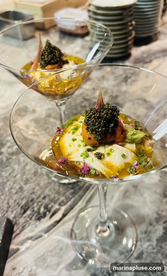 Two martini glasses filled with a delicate appetizer. The appetizer features a creamy white base topped with a generous portion of black caviar, a small piece of orange sea urchin, and garnished with chives and edible flowers. The dish is presented in a rich, golden broth. In the background, stacks of plates and kitchen equipment are visible.
