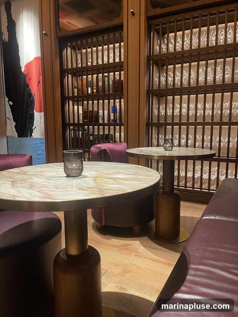 The image shows the interior of a bar or lounge area. There are several round tables with marble-patterned tops and wooden bases, surrounded by plush seating in shades of burgundy and purple. Behind the tables, tall wooden shelves are filled with bottles and glassware, illuminated by soft lighting. A large, abstract artwork with red and black elements hangs on the left wall. The overall ambiance is sophisticated and dimly lit.