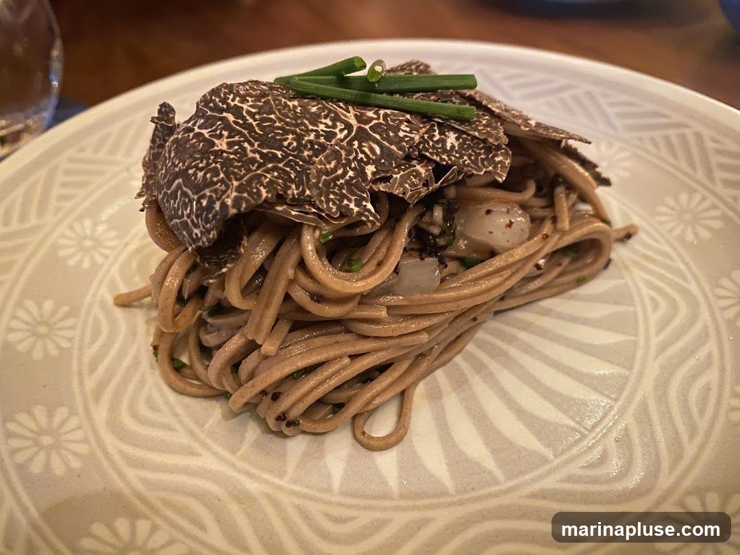 A close-up shot of a plate of pasta topped with truffle shavings and chives. The pasta appears to be whole wheat or buckwheat noodles, mixed with small pieces of seafood and herbs. The plate has a decorative pattern.