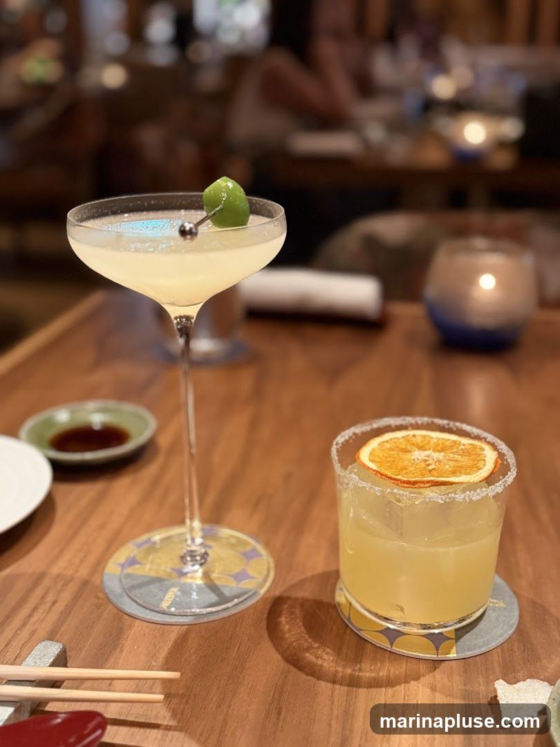 Two cocktails are on a wooden table. One is in a coupe glass with a lime garnish. The other is in a rocks glass with a dried orange slice and a salt rim. There are chopsticks, a small dish of sauce, and a candle in the background.