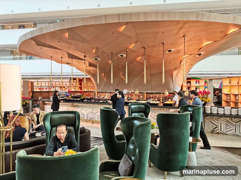 A contemporary bar and lounge area with plush green seating and a distinctive, undulating wooden ceiling. Patrons are seated at tables and at the bar, where staff in chef's attire are visible. The bar itself is well-stocked with bottles and displays food items. The overall ambiance is sophisticated and inviting.