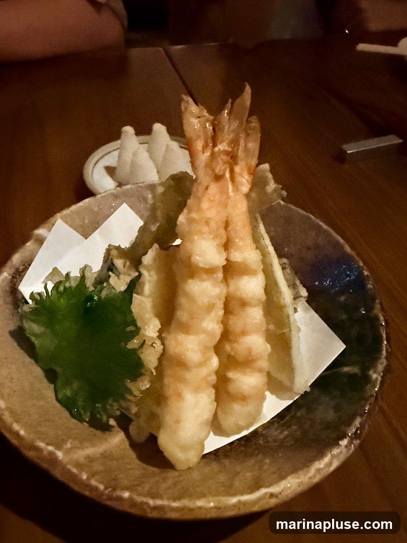 A close-up shot of shrimp tempura served in a rustic bowl on a wooden table. The tempura is golden brown and crispy, with two large shrimp prominently displayed. A green leaf garnish and a small dish of white condiments are also visible.