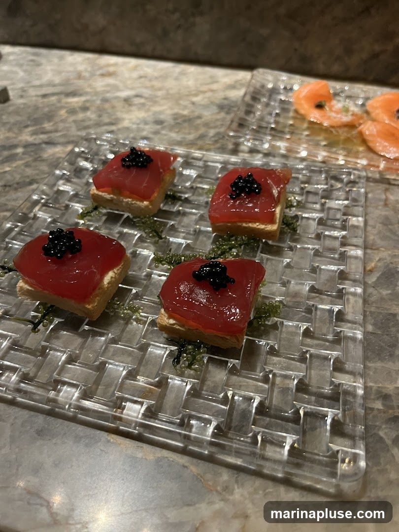 Four square appetizers topped with tuna and caviar are arranged on a clear, woven plate. In the background, thinly sliced salmon is served on another clear plate. The appetizers are garnished with green sea grapes.