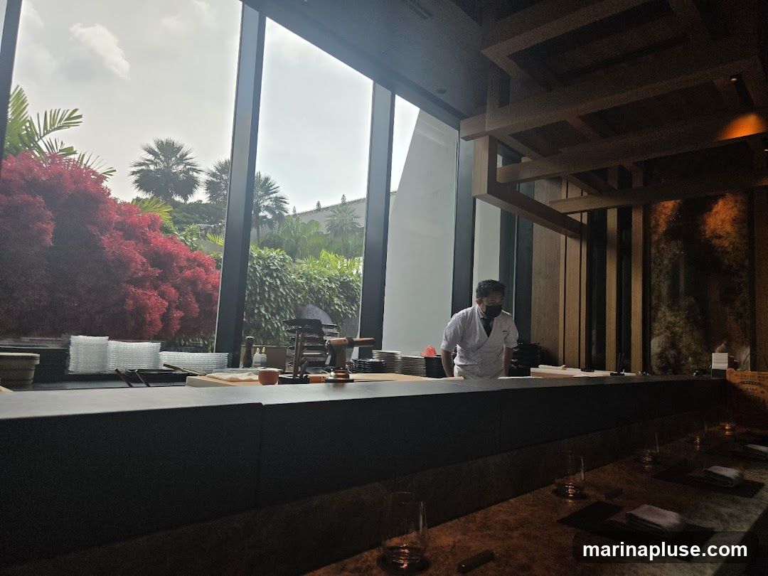 A chef in a white uniform and mask stands behind a sushi bar, preparing food. Large windows offer a view of lush greenery and palm trees outside. The interior features a modern, minimalist design with wooden accents and warm lighting. The sushi bar is set with plates, glasses, and napkins, ready for customers.