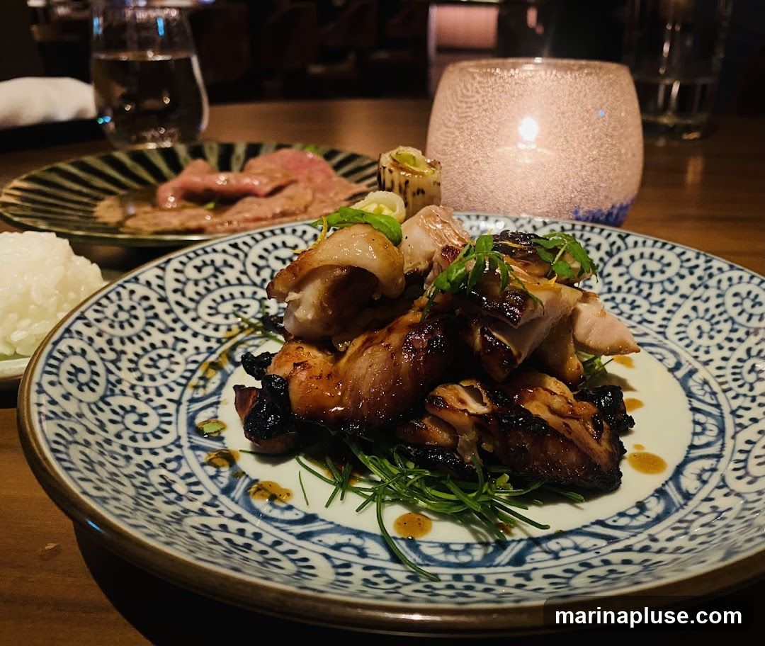 A close-up shot of a plate of grilled chicken with a side of rice and thinly sliced beef. The chicken is glazed and appears to be seasoned with herbs. The dish is served on a decorative blue and white patterned plate. In the background, a lit candle in a glass holder and glasses of water are visible, suggesting a restaurant setting.