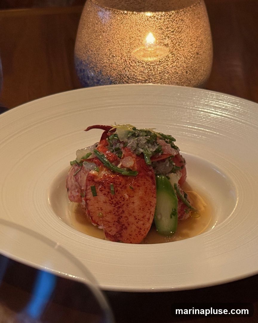 A close-up shot of a gourmet lobster dish served in a white bowl. The lobster meat is bright red and appears to be chilled, topped with finely chopped herbs, possibly chives, and a light dressing. Accompanying the lobster are pieces of green asparagus. The dish is presented with a warm, ambient glow from a decorative candle holder in the background, creating an intimate dining atmosphere. A wine glass is partially visible in the foreground, suggesting a fine dining experience.