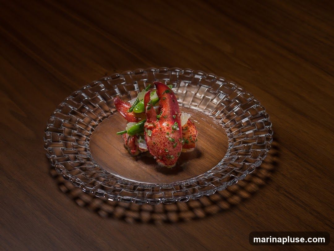 A close-up shot of a lobster appetizer served on a clear, woven glass plate. The dish features pieces of lobster claw and tail, mixed with finely chopped green herbs and possibly some citrus zest. The plate is set on a dark wooden surface, with dramatic shadows cast by the plate's intricate design.