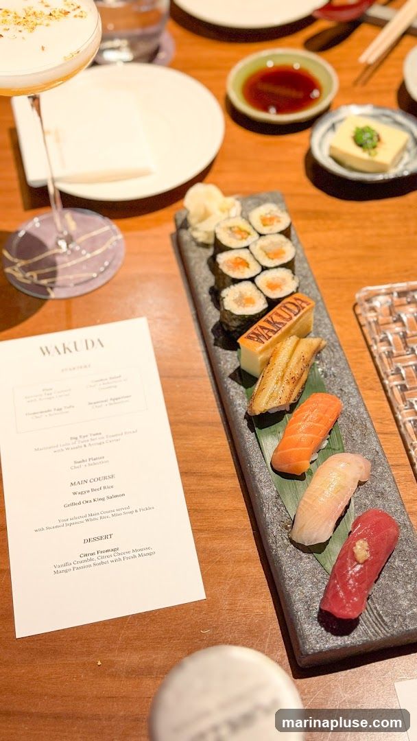 A close-up view of a sushi platter and a menu from Wakuda. The platter features various sushi pieces, including tuna, salmon, and eel, along with a piece of tamagoyaki branded with the restaurant's name. A cocktail with gold flakes is visible in the foreground. The menu lists starters, main courses like Wagyu Beef and Grilled Ora King Salmon, and a dessert called Citrus Fromage.