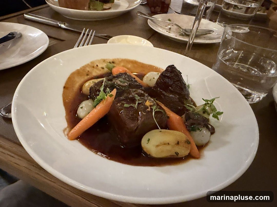 A close-up shot of a white plate filled with a braised beef dish. The beef is dark and tender, served in a rich brown sauce. Accompanying the beef are small, whole carrots and pearl onions, along with some small potatoes. A garnish of fresh green herbs is sprinkled on top. The plate is set on a dark wooden table, with other plates and glasses visible in the background, suggesting a dining setting.