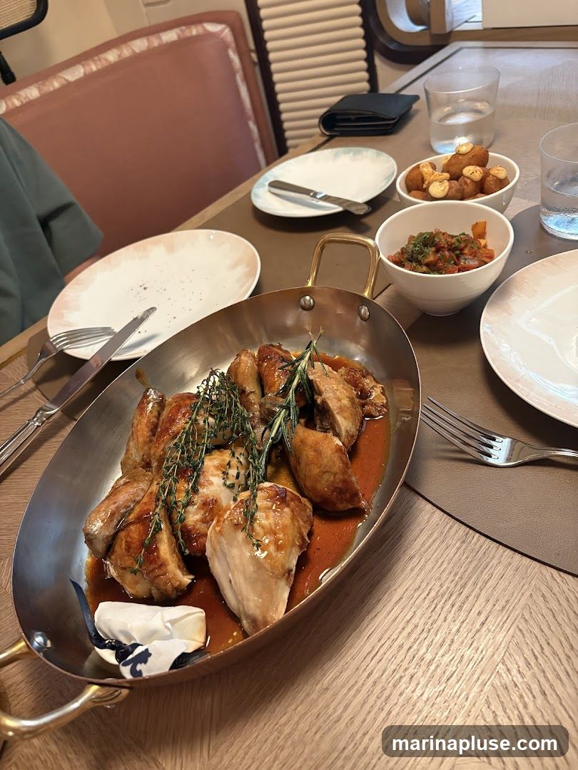 A whole roasted chicken, carved and served in a copper pan with a rich sauce and garnished with fresh rosemary and thyme. Two small bowls of appetizers, one with fried croquettes and the other with a tomato-based relish, are visible on the table, along with plates, cutlery, and glasses of water. The setting appears to be a restaurant dining area with upholstered seating in the background.