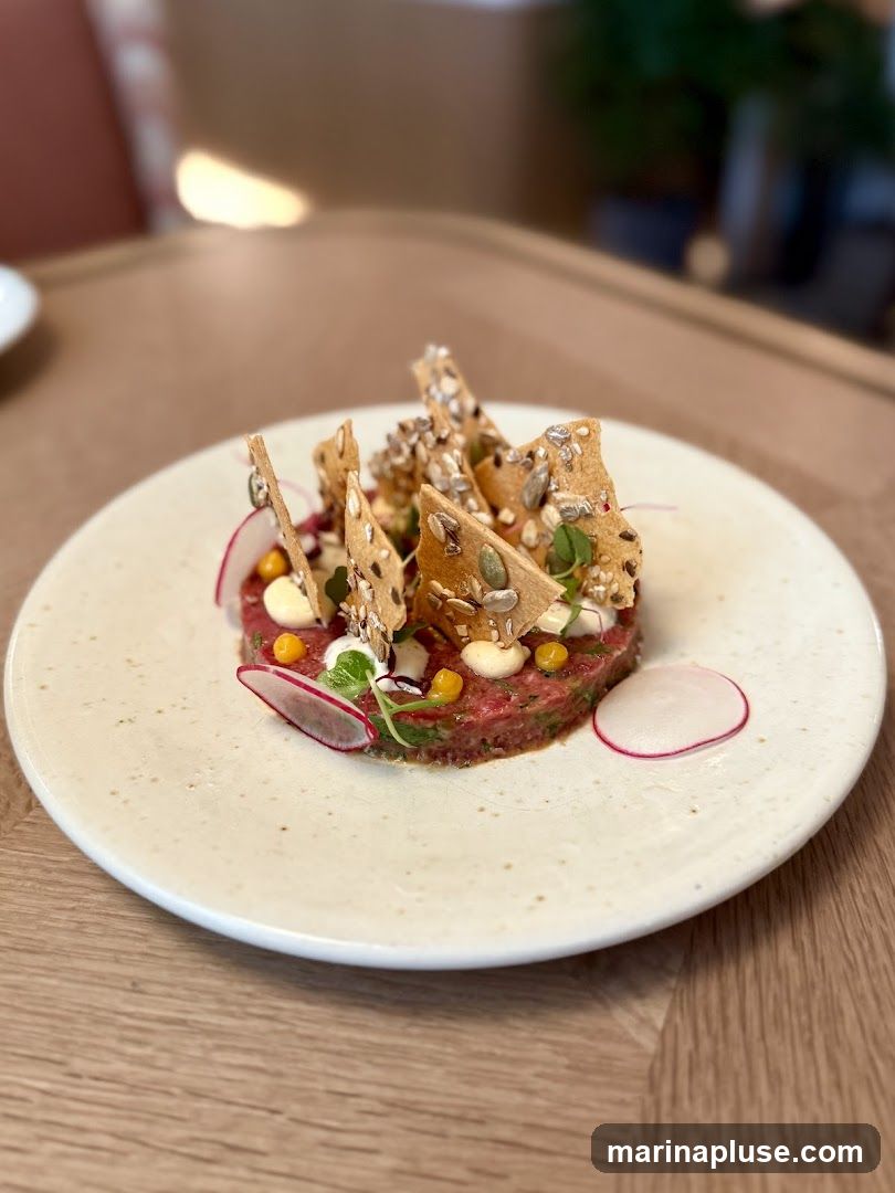 A close-up shot of a beef tartare dish served on a white speckled plate. The tartare is topped with various seeds, herbs, and crispy crackers. Thinly sliced radishes and small yellow spheres are also visible. The dish is presented on a wooden table, with a blurred background suggesting an indoor dining setting.