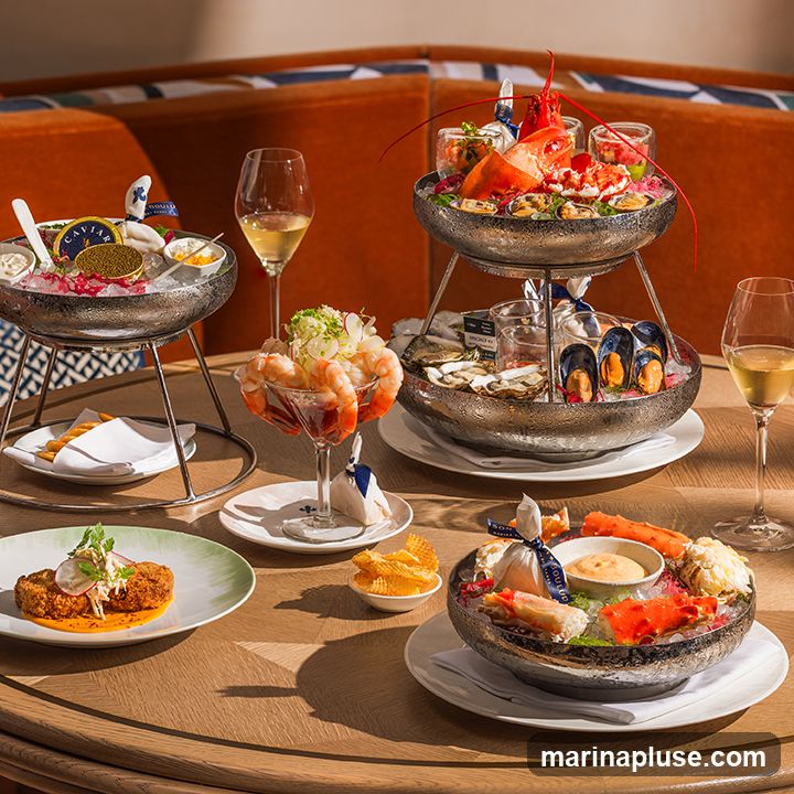 A lavish seafood platter is presented on a wooden table, featuring a tiered display of fresh oysters, mussels, lobster, and crab legs. Accompaniments include cocktail sauce, caviar, and a fried dish with garnish. Two glasses of white wine and a small bowl of potato chips are also visible. The background shows orange upholstered seating.
