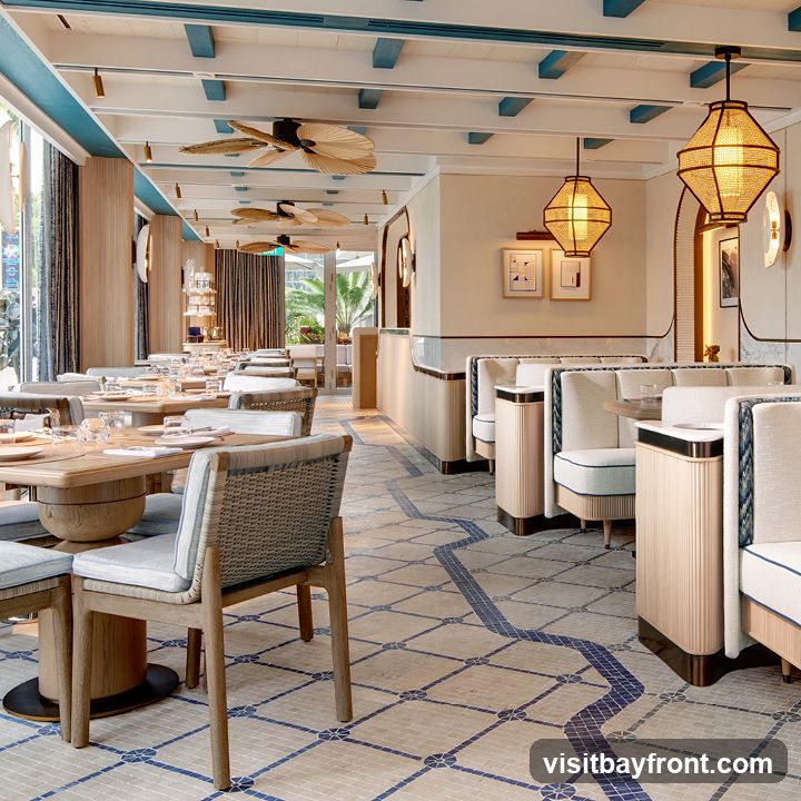 A well-lit and elegantly designed restaurant interior with a focus on natural materials and a coastal-inspired aesthetic. The space features wooden tables set with white tableware, comfortable seating with upholstered cushions, and woven chairs. The ceiling is adorned with exposed wooden beams and decorative fans, while unique pendant lights cast a warm glow. The floor is tiled with a mosaic pattern in beige and blue. Booths line one side of the dining area, offering a more intimate seating option. Large windows allow natural light to flood the space, and glimpses of greenery can be seen outside.