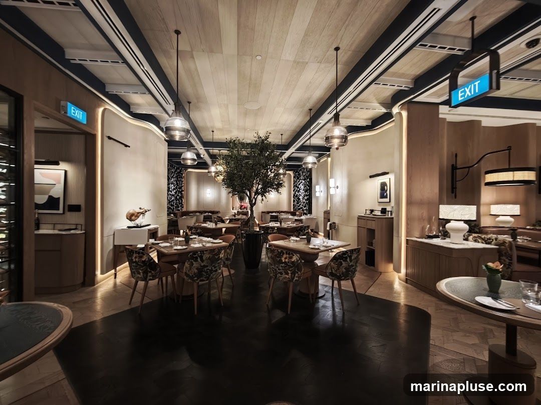 The interior of a modern restaurant with elegant decor. Tables are set with chairs featuring floral upholstery. A large potted tree stands in the center of the dining area. Ambient lighting is provided by pendant lamps and wall sconces. Exit signs are visible.