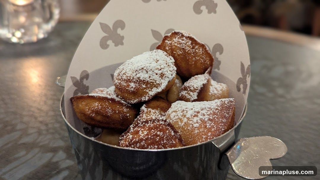 A close-up shot of beignets, a type of fried dough pastry, dusted generously with powdered sugar. They are served in a metal container lined with decorative paper featuring fleur-de-lis patterns. The background is blurred, showing a reflective surface and a hint of a candle holder.