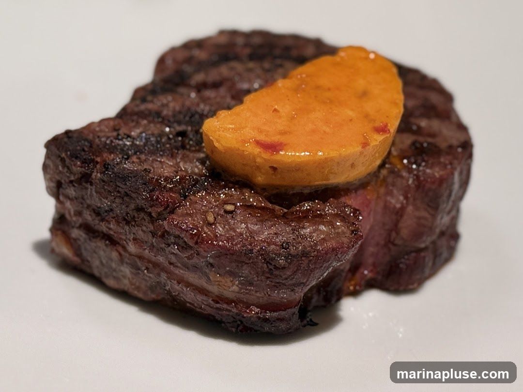 A close-up shot of a grilled steak, topped with a slice of orange-colored food, possibly a vegetable puree or butter. The steak has visible grill marks and appears to be cooked to medium-rare. The background is a plain white surface.