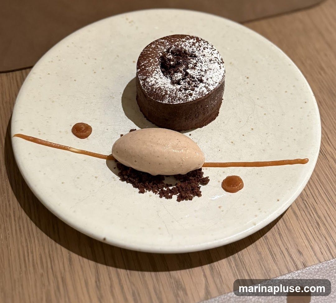 A close-up shot of a chocolate lava cake dusted with powdered sugar, served with a scoop of light brown ice cream and chocolate crumbs on a white plate. A thin line of caramel sauce and two dollops of caramel sauce adorn the plate. The plate is set on a wooden table.