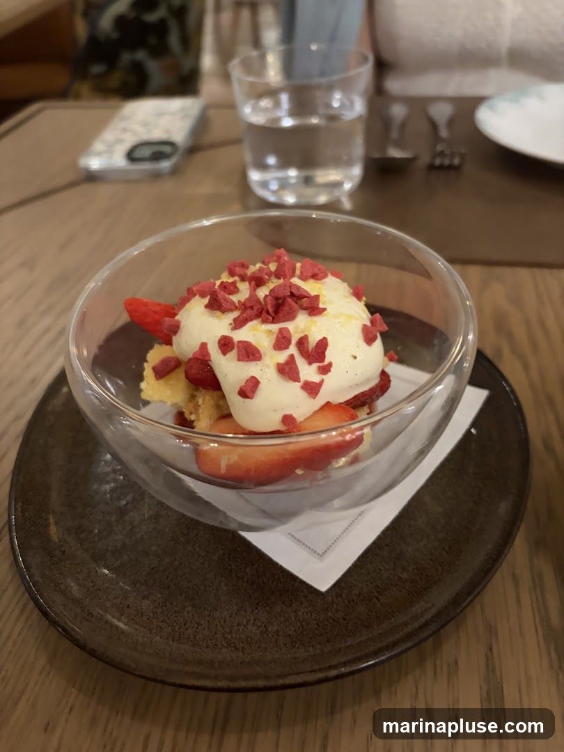 A close-up shot of a dessert served in a glass bowl. The dessert appears to be a layered cake or crumble with fresh strawberries and a dollop of cream on top, garnished with red sprinkles. The bowl sits on a dark plate on a wooden table. In the background, a glass of water and a smartphone are visible.