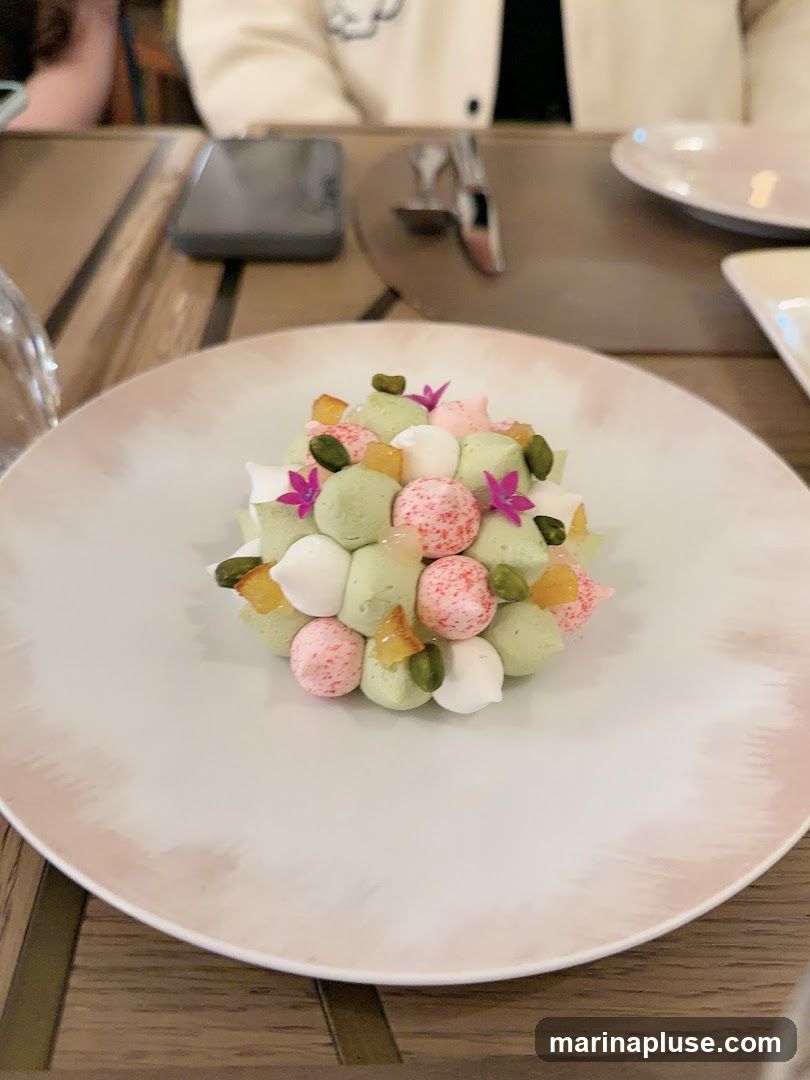 A close-up shot of a dessert served on a white plate with a pink rim. The dessert is a colorful arrangement of small, round meringue-like pieces in shades of green, white, and pink, some speckled with red. It is garnished with chopped pistachios and small pieces of candied fruit, as well as delicate purple edible flowers. The plate sits on a wooden table, and a person's arm and shoulder are visible in the background, along with a phone and cutlery.