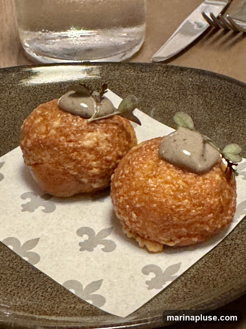 Two golden-brown, spherical appetizers are presented on a dark plate, each topped with a dollop of greyish-brown spread and garnished with microgreens. The appetizers are placed on a white paper liner featuring a subtle fleur-de-lis pattern. A glass of water and silverware are visible in the background, suggesting a dining setting.