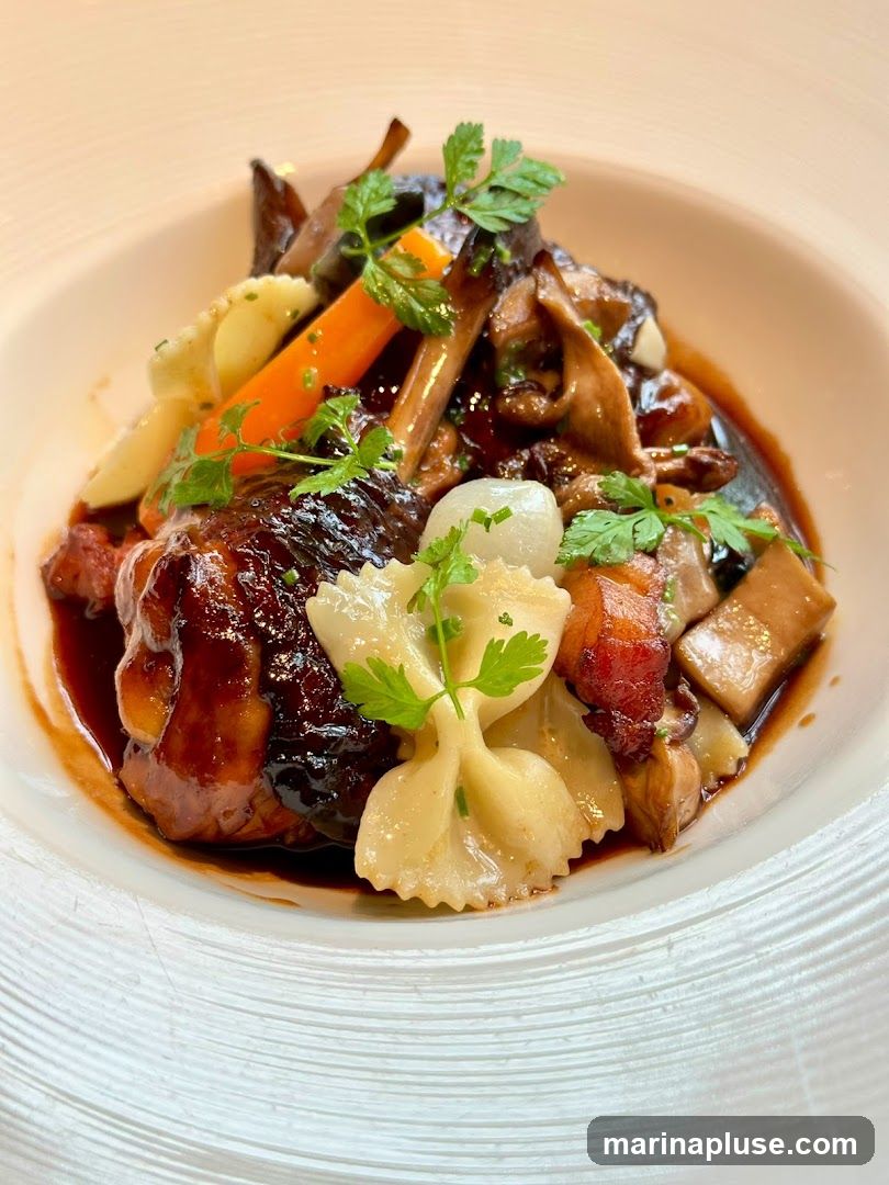 A close-up shot of a rich, dark braised dish served in a white bowl. The dish features tender meat, possibly beef or duck, coated in a glossy sauce. It is accompanied by farfalle pasta, chunks of carrots, mushrooms, and pieces of bacon. Fresh parsley is used as a garnish. The bowl has a textured rim.