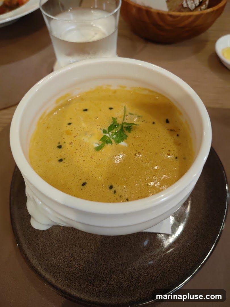 A close-up shot of a bowl of creamy, orange-colored soup, garnished with a sprig of parsley and black specks. The soup is served in a white bowl with decorative detailing on its base, which sits on a dark brown saucer. In the background, a glass of water and a wooden bowl of bread are visible.