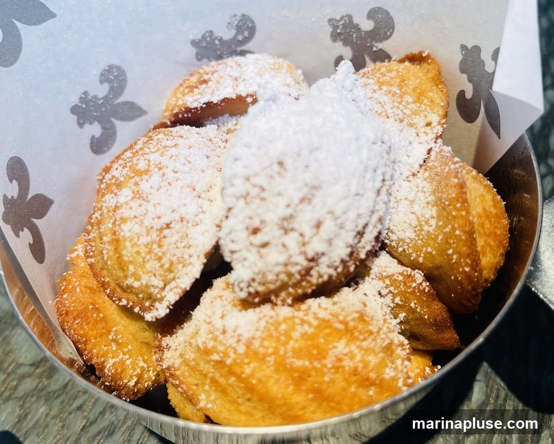 A close-up shot of beignets, a type of fried dough pastry, generously dusted with powdered sugar. They are served in a metallic bowl lined with white paper featuring a fleur-de-lis pattern. The beignets appear golden brown and fluffy.