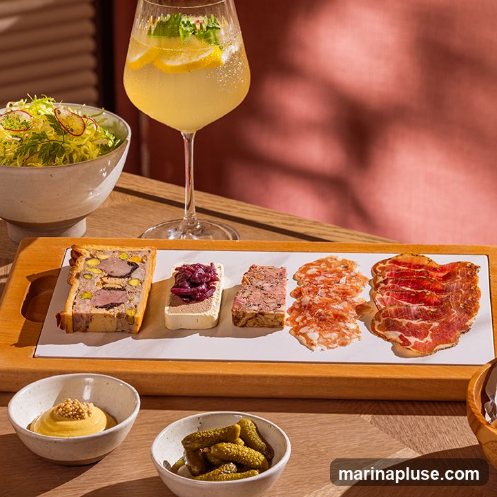 A charcuterie board featuring several types of pâté and cured meats, served with small bowls of mustard and cornichons. A glass of lemonade with lemon slices and mint is in the background, along with a side salad.