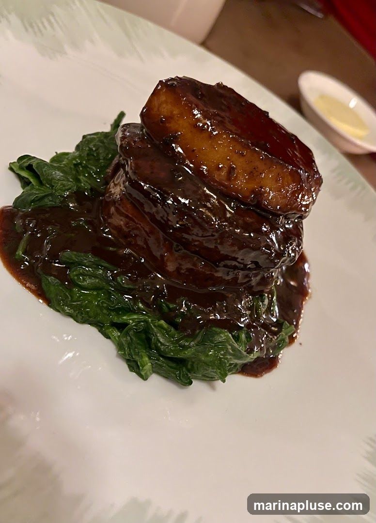 A close-up shot of a dish featuring braised pork belly stacked on a bed of wilted spinach. The pork belly is coated in a rich, dark glaze and appears tender. A small dish of butter or sauce is visible in the blurred background.