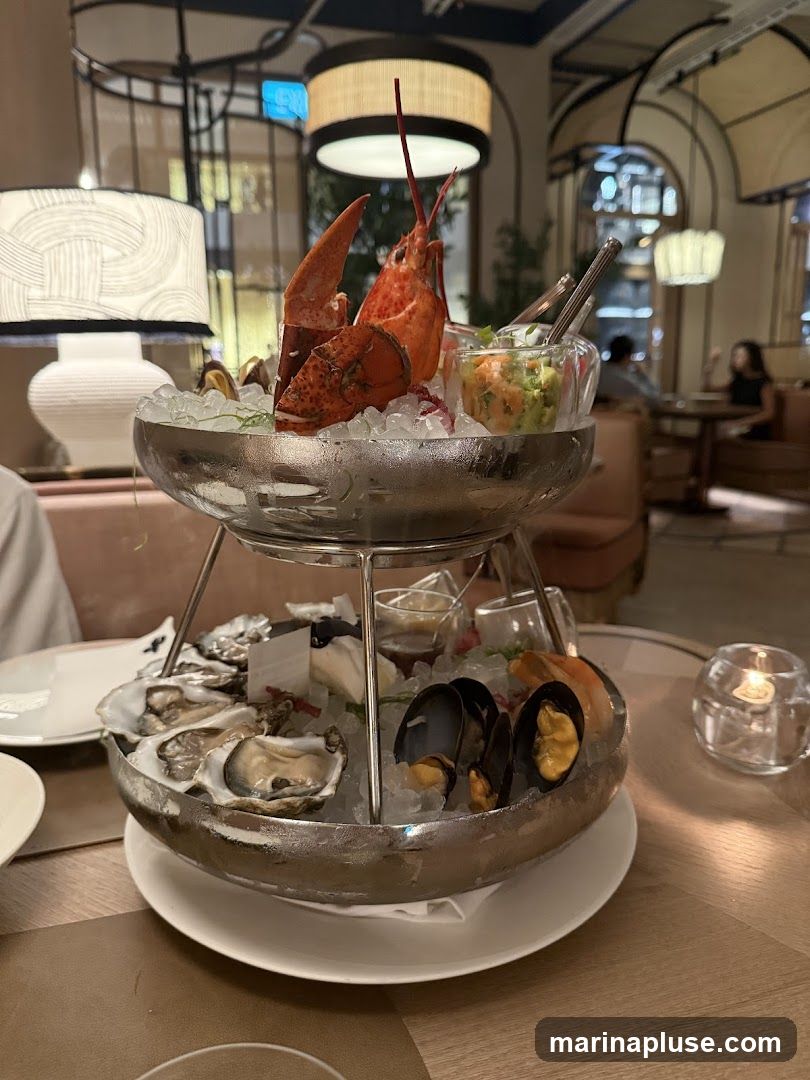 A tiered seafood platter is presented on a table in a restaurant. The top tier features a whole lobster, a small bowl of what appears to be a salsa or relish, and some mussels. The bottom tier holds a generous portion of oysters, mussels, and a shrimp. The seafood is arranged on a bed of crushed ice. In the background, the restaurant's interior is visible, with elegant lighting fixtures, upholstered seating, and other diners.