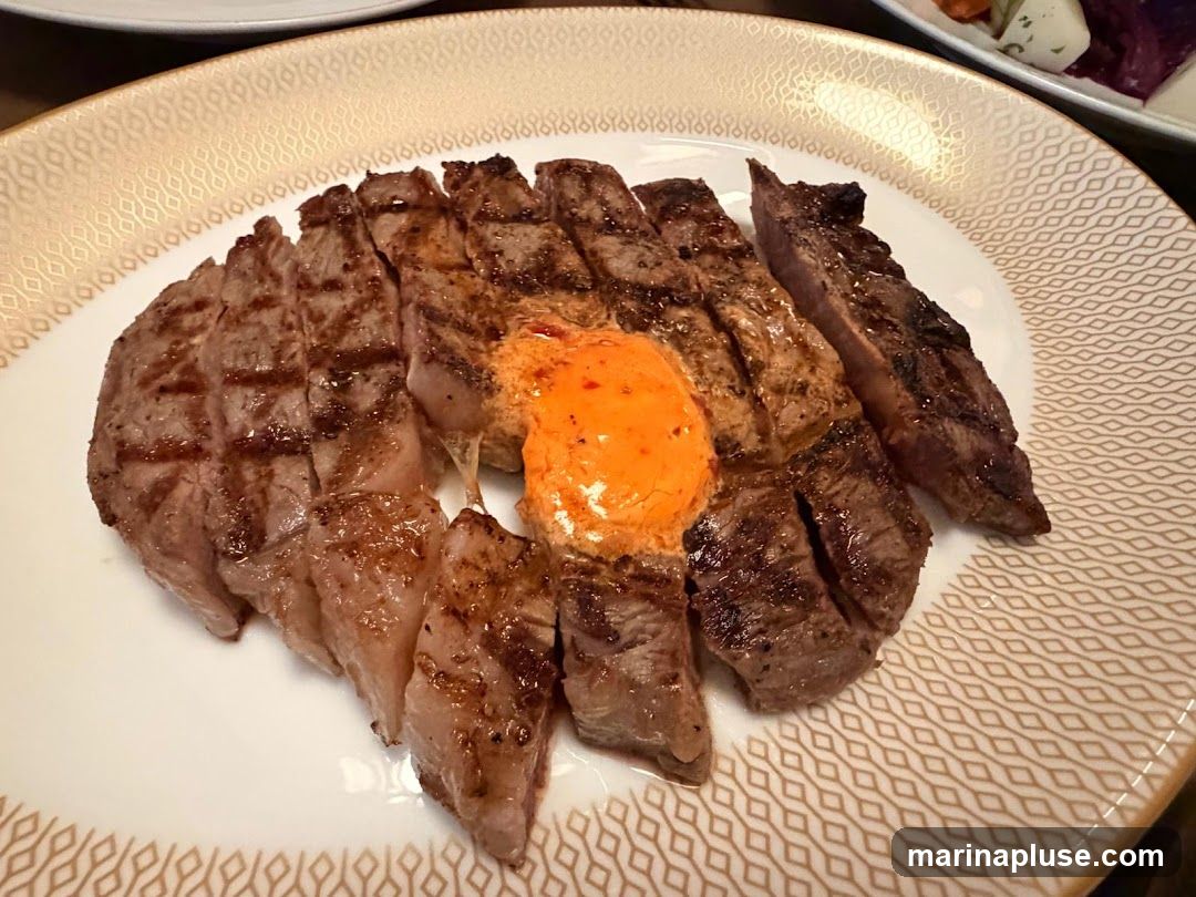 A close-up shot of a sliced steak served on a white plate with a decorative gold trim. The steak is grilled, showing distinct grill marks, and is topped with a dollop of bright orange butter or sauce. In the background, out of focus, is a salad with purple and white ingredients.