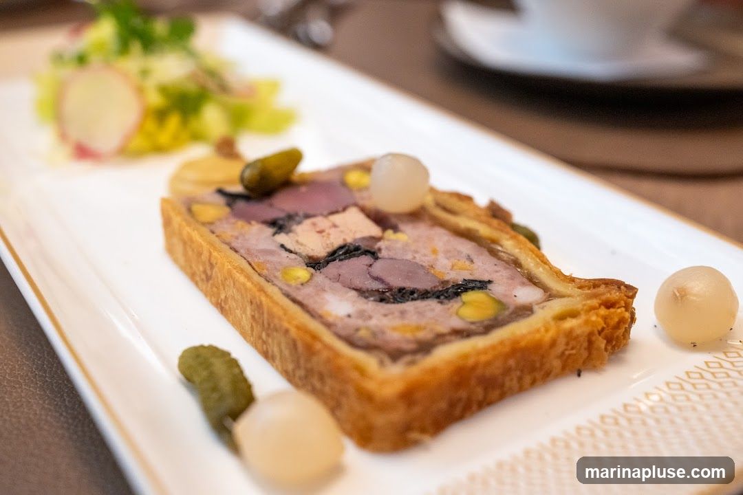 A slice of terrine is served on a white rectangular plate with a gold trim. The terrine appears to be made of various meats and includes pistachios and what looks like foie gras. It is garnished with cornichons and pearl onions. A small salad with lettuce and radish slices is visible in the background.