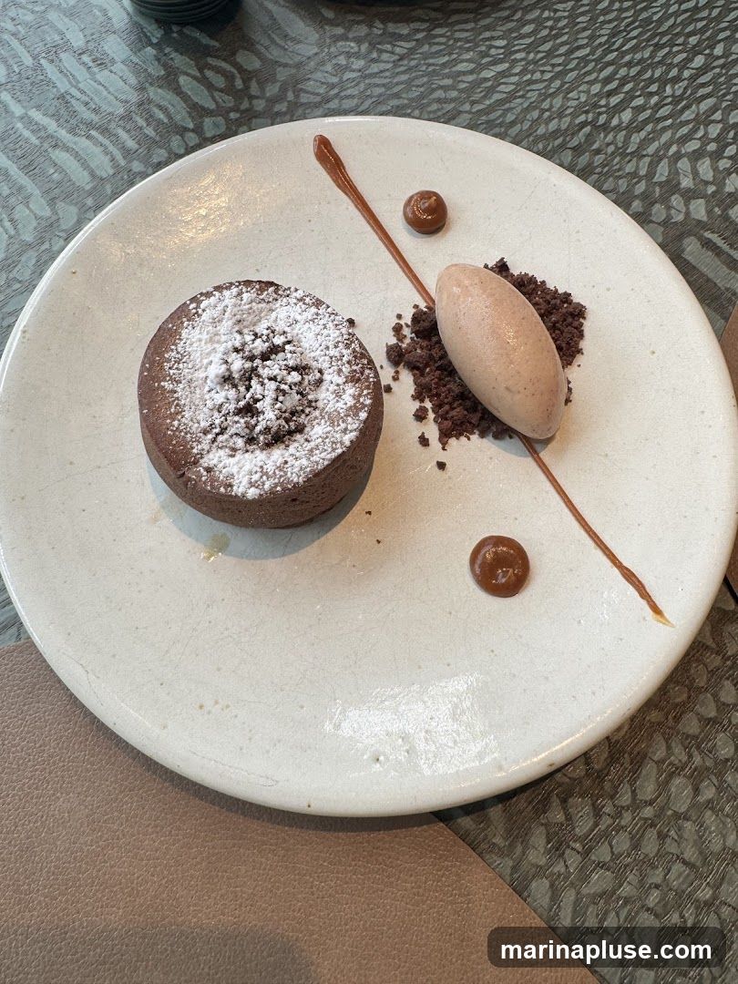 A close-up shot of a chocolate lava cake dusted with powdered sugar, served on a white plate. Alongside the cake is a scoop of light brown ice cream, a pile of chocolate crumbles, and a drizzle of chocolate sauce with two small dollops of caramel. The plate is set on a textured surface, possibly a table.