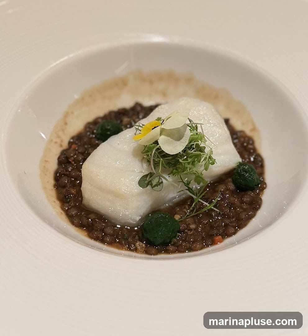 A white fish fillet is served on a bed of dark lentils in a white bowl. The dish is garnished with microgreens, edible flowers, and small green spheres.