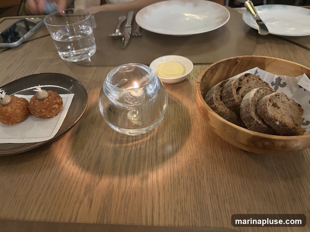 A table setting with two appetizers on a dark plate, a small dish of butter, a bowl of sliced bread, a glass of water, and a lit candle in a glass holder. Plates and cutlery are visible in the background.