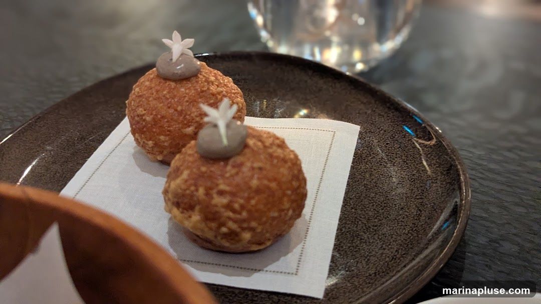 Two small, golden-brown, spherical appetizers are presented on a dark, textured plate, each topped with a dollop of grey cream and a delicate white flower. They are placed on a white napkin with a stitched border. A blurred glass and a wooden bowl are visible in the background.