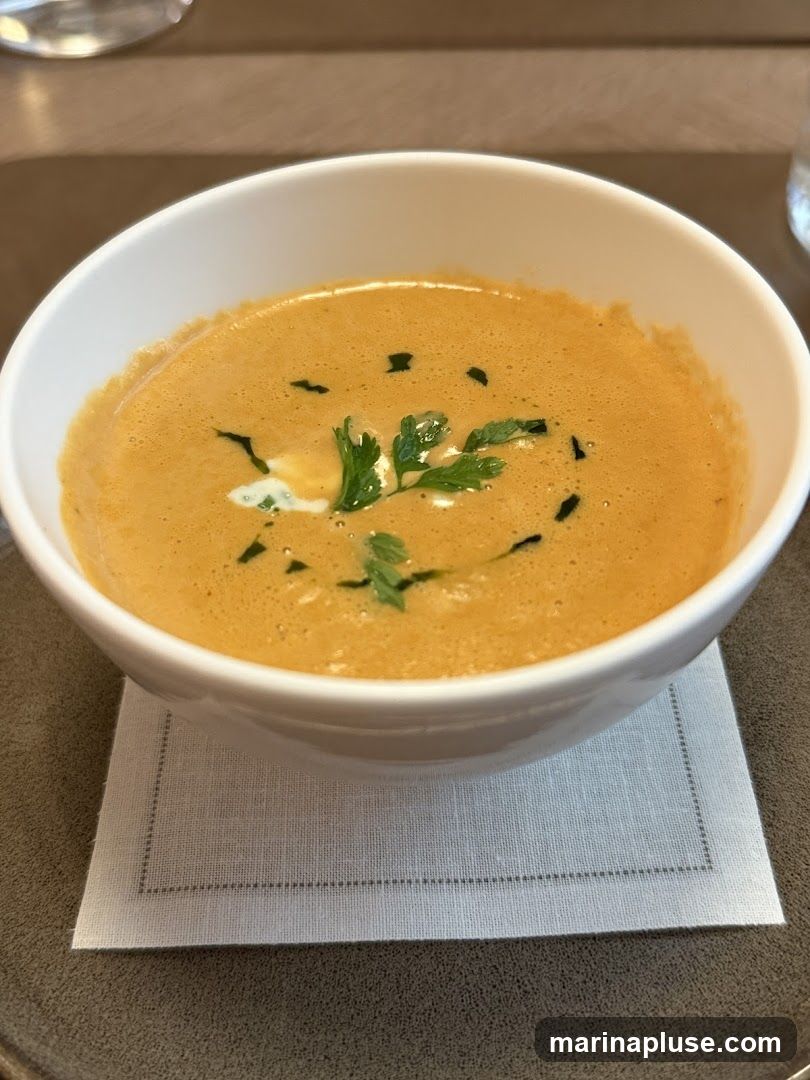 A close-up shot of a bowl of creamy, orange-hued soup. The soup is garnished with a swirl of white cream and fresh green herbs, possibly parsley. It is served in a white bowl placed on a light grey napkin, which rests on a dark grey placemat. The background is blurred, suggesting an indoor dining setting.