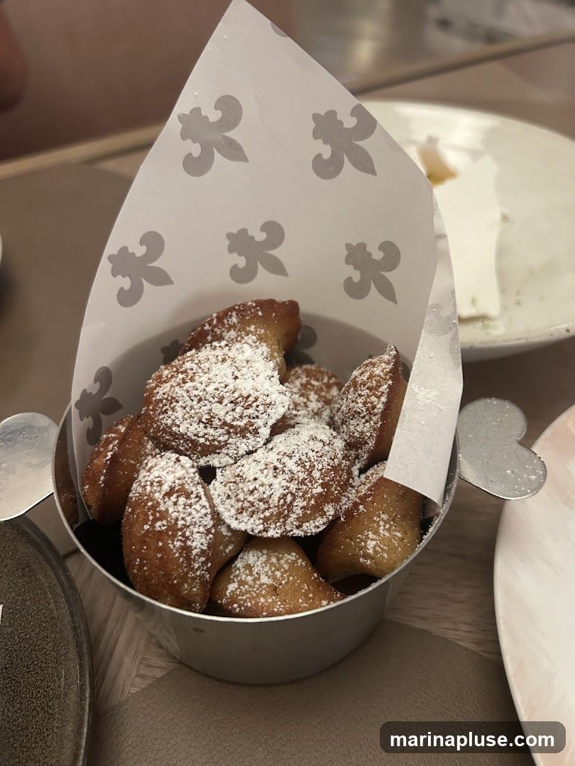 A close-up shot of madeleines dusted with powdered sugar, served in a metal container lined with decorative paper. In the background, a white dessert with a yellow topping is visible on a plate.